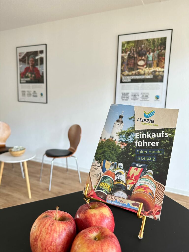 Im Vordergrund sind drei rote Äpfel und der aufgestellte Fairtrade Einkaufsführer zu sehen. Im Hintergrund sind zwei Poster der Ausstellung „Leipzig handelt fair“ zu sehen.